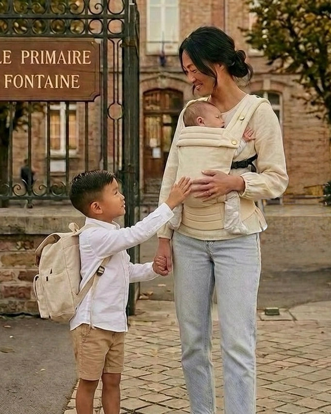 Parent portant bébé en porte‑bébé physiologique beige, accompagné d’un enfant marchant devant une école — portage pratique pour trajets quotidiens